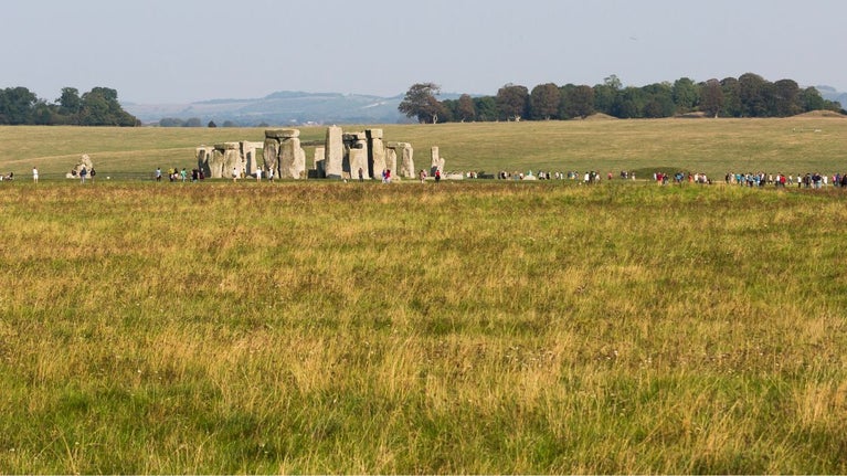 A wide landscape shot across a chalk grassland field with the Stonehenge monument and visitors visible in the background